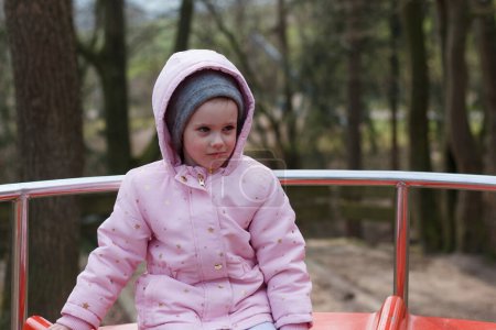 Une petite fille dans un capot et un chapeau chaud monte seul sur un carrousel dans un parc d'hiver
