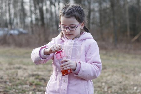 Lächelndes kleines Mädchen in Brille spielt mit Seifenblasen in einem schneelosen Winterpark