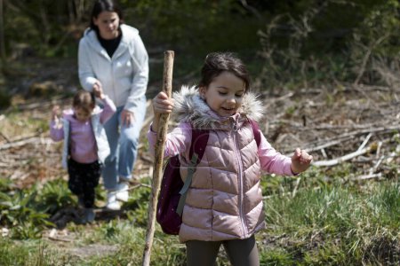 A little girl with a stick and a staff walks through the forest. Mom and sister in the background
