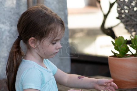 A little sad girl with a ponytail stands by a pot with an indoor flower on the terrace