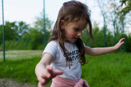 Téléchargez les photos : Belle petite fille maternelle dans un T-shirt blanc souriant marche sur une corde sur une aire de jeux d'été - en image libre de droit
