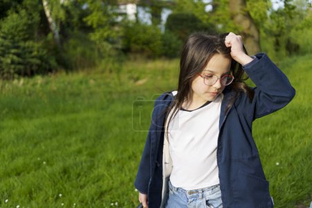 A little girl in glasses, a T-shirt and a jacket scratches her head while standing on a spring street