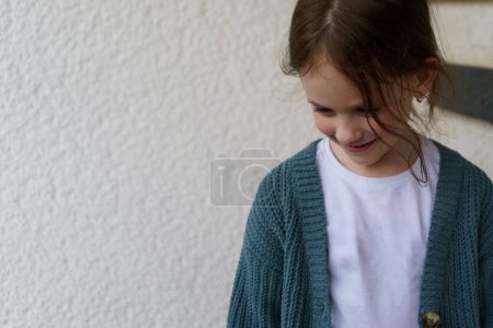 Una niña sonriente avergonzada con una camiseta y una chaqueta se encuentra cerca de la entrada de la casa