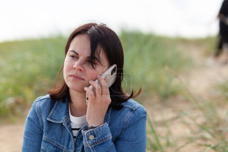 Hermosa chica morena hablando por teléfono mientras está sentada en una playa de dunas de verano con una chaqueta de mezclilla