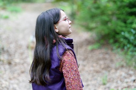 Una pequeña hermosa chica de pelo largo en gafas y un chaleco mira hacia arriba mientras está de pie en el bosque
