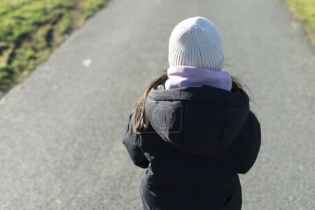 A little girl, warmly dressed, walks along a village asphalt road, close-up view from behind