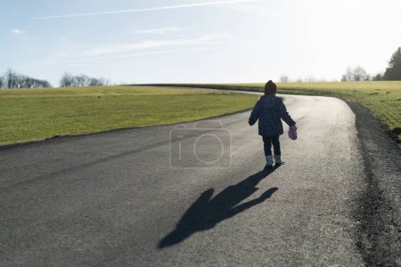 Beautiful winter photo of a little warmly dressed girl with a shadow on a rural asphalt winter road without snow, rear view