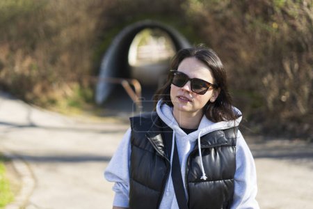 A cute dark-haired girl in sunglasses, a vest and a hoodie stands smiling in a spring park