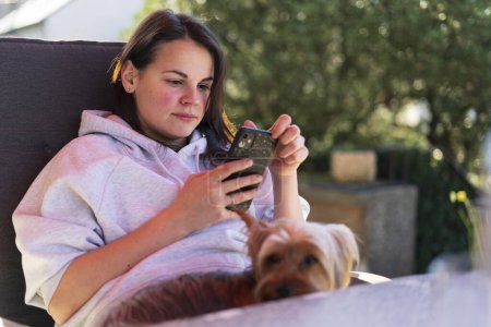 Une belle jeune fille en sweat à capuche léger lit sur son téléphone portable alors qu'elle est assise à une table sur la terrasse de sa maison. Concept de technologie cellulaire à l'extérieur