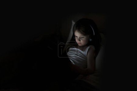 Una pequeña chica hermosa con auriculares se encuentra con una tableta en una habitación oscura. El concepto de la vida digital moderna y la infancia