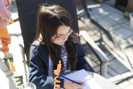 A little girl with glasses sits at a table in the backyard and does her homework. Educational concept