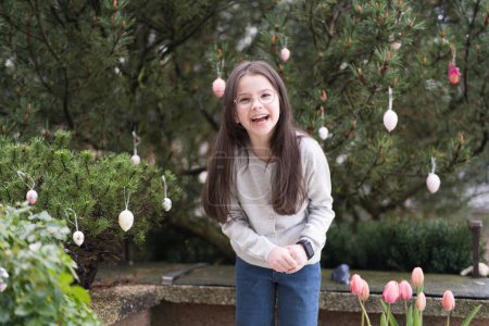 A little long-haired girl with glasses stands smiling near an Easter tree.