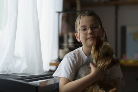 Ein kleines hübsches Teenager-Mädchen mit Brille und Frisur sitzt mit einem Hund am Klavier. Im Hintergrund verschwommenes Kinderzimmer. Konzept von Erziehung, Kindheit und Freundschaft mit einem Haustier