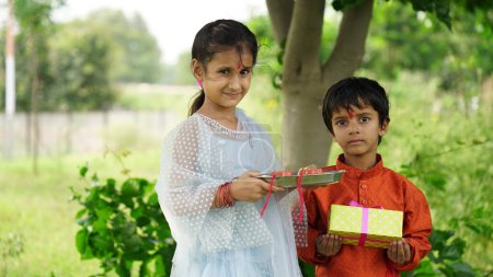 Téléchargez les photos : Jolie petite sœur indienne attachant Rakhi au poignet de son petit frère et échangeant cadeaux et bonbons sur le festival Raksha Bandhan ou Bhai Dooj - en image libre de droit