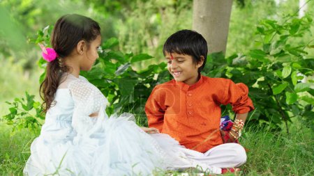 Téléchargez les photos : Jolie petite sœur indienne attachant Rakhi au poignet de son petit frère et échangeant cadeaux et bonbons sur le festival Raksha Bandhan ou Bhai Dooj, assise sur fond de nature - en image libre de droit