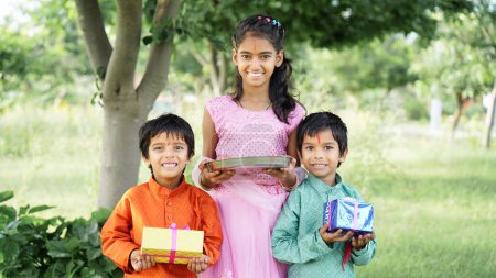 Téléchargez les photos : Frères hindous et sœur en tenue ethnique tenant des bonbons indiens et des coffrets-cadeaux à l'occasion du festival Raksha Bandhan - en image libre de droit