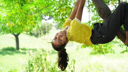 Foto de Linda niña juguetonamente cuelga de una rama de árbol. Hora de verano, calor, infancia. Retrato divertido con cabello desaliñado - Imagen libre de derechos
