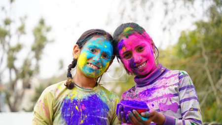 Photo for Happy Indian friends or kids celebrating the Hindu festival of Holi by applying colors to each other, best friends. Female siblings holding a plate with different shades of Gulal - Royalty Free Image