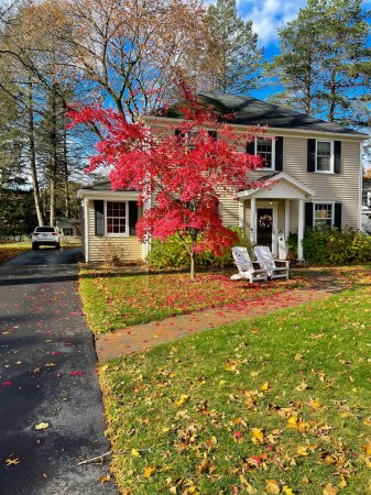 Lange Autofahrt zu einem zweistöckigen Haus mit bunten roten Ahornblättern und weißen Adirondack-Stühlen auf der Veranda in einem Vorort von Rochester, New York, USA. Sitzgelegenheiten und Wohnraum im Vorgarten im Herbst