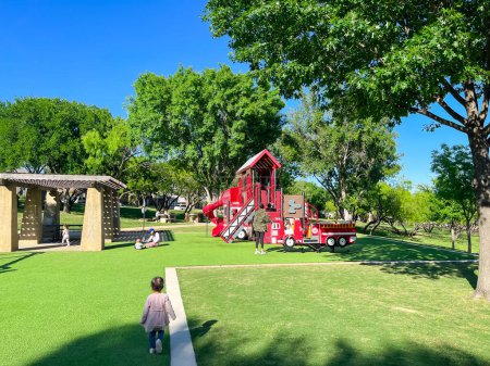 Photo for Rear view unidentified kids walking, running at firetruck themed playground with green artificial grass turf carpet, tall trees clear blue sky in Carrollton, suburbs Dallas, Texas, USA. Suburban park - Royalty Free Image