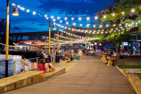 Photo for Blurry people hanging out at waterfront boardwalk string lights, beach chairs, patio restaurant dining tables sunset blue hour with elevated highway, skylines near Dallas, Texas, USA. Lakeside eatery - Royalty Free Image