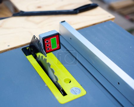 Photo for Wooden workbench with brand new table saw has steel blade attaches to digital level box inclinometer showing 0-degree angle calibration and plastic push stick in background. Protractor bevel gauge - Royalty Free Image