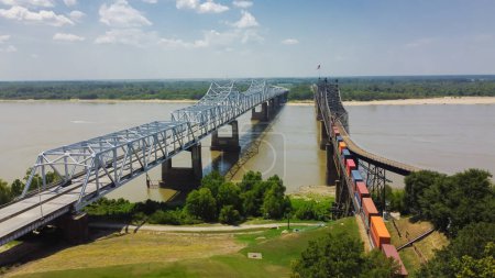 Téléchargez les photos : Pont de Vicksburg et Vieux Pont de Vicksburg avec train de marchandises traversant la rivière ferme ferroviaire entièrement en acier transportant l'Interstate 20, U.S. Route 80 à travers le ciel nuageux ensoleillé du fleuve Mississippi. Vue aérienne - en image libre de droit