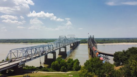 Téléchargez les photos : Pont de Vicksburg et Vieux Pont de Vicksburg avec train de marchandises traversant la rivière ferme ferroviaire entièrement en acier transportant l'Interstate 20, U.S. Route 80 à travers le ciel nuageux ensoleillé du fleuve Mississippi. Vue aérienne - en image libre de droit