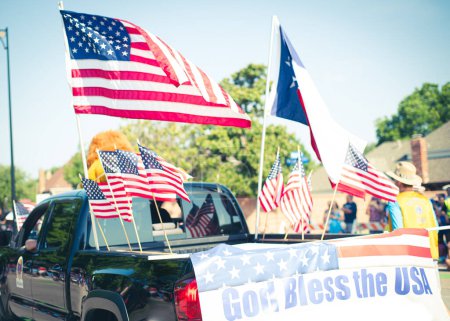 Téléchargez les photos : Que Dieu bénisse la bannière américaine arrière camionnette moderne dense du drapeau américain sur le lit de cargaison conduite sur la petite ville de rue résidentielle Quatrième défilé de Juillet, Dallas, Texas, États-Unis personnes floues. Jour de l'indépendance - en image libre de droit
