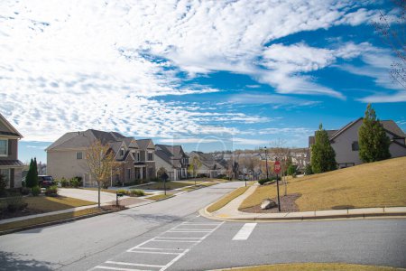 Dreispurige Straßenkreuzung mit Stoppschild, zweistöckige Neubausiedlung entlang der Straße, die in der Nähe von Atlanta, Georgia, USA, einen steilen Hügel hinunter führt, gepflegte Landschaft. Sonniger wolkenblauer Himmel