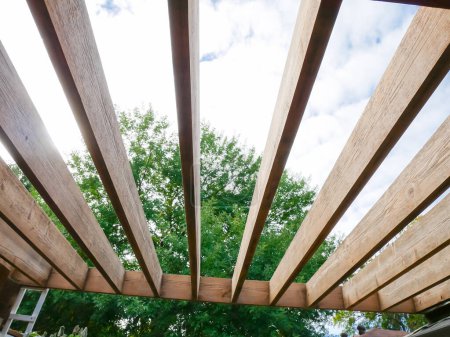 Recently sanded pergola roof rafter with large tree in background, ready for re-stain wooden beams overhang post timber outdoor structure backyard garden Dallas, Texas, USA, natural wood grain. DYI