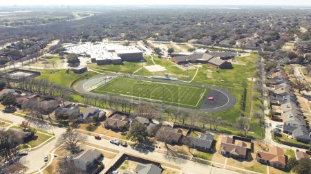 Middle school football stadium with artificial turf, yard markings, track and field situated in upscale residential neighborhood outside Dallas, Texas, lake and highway in distance background. Aerial