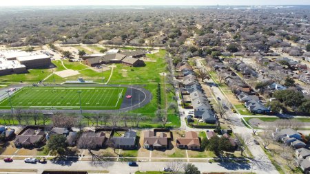 Residential neighborhood and school district with row of single family homes, football stadium artificial turf, yard markings, track and field, subdivision urban sprawl in Dallas, Texas, USA. Aerial
