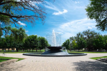 Tall water fountain at brick campus quad courtyard in Waco, Texas, lush green tree, grassy front yard, row of historic buildings background, summer sunny cloud blue sky, education and landscaping concept. USA