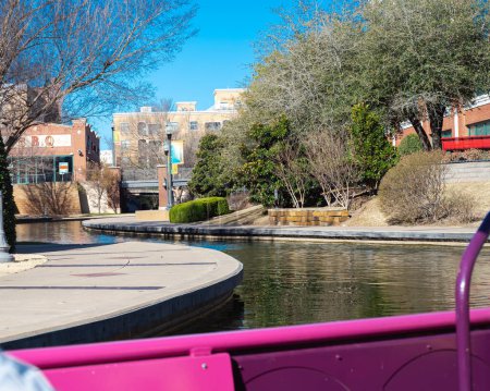Water taxi cruises along canal with riverside buildings, hotels, restaurants in Bricktown, Entertainment District, Oklahoma City, wintertime bare trees, travel destination and tourist attractions. USA