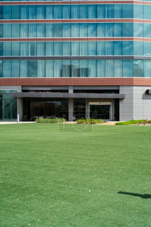 Facade entrance of modern hotel with large artificial grass courtyard, glass wall with blackout curtains, multistory building exterior in downtown Irving, Texas, urban travel lifestyle. USA