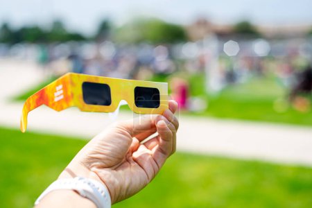 Asian hand wear smart watch holding paper solar eclipse with blurry crowd people watching totality show in Dallas, Texas, April 8, scratch resistant polymer lenses filter out harmful ultraviolet. USA