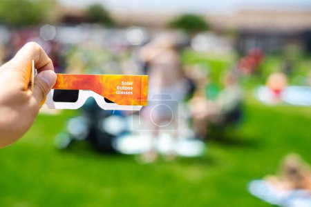 Side view of paper optics solar eclipse glasses scratch resistant polymer lenses filter harmful ultraviolet, infrared ray, blurry crow people on grassy yard watching totality show, Dallas, Texas. USA