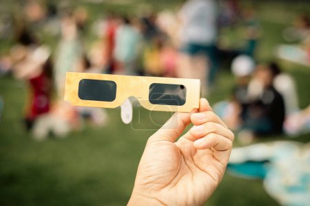 Hand holding paper solar eclipse with blurry crowd people watching totality show picnic yard, Dallas, Texas, April 8, scratch resistant polymer lenses filter out harmful ultraviolet, infrared ray. USA