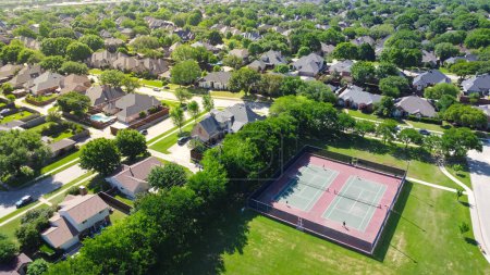 Community tennis courts with chain link fence and players in upscale residential neighborhood with large two-story single-family house, swimming pool, fenced backyard, suburb Dallas, TX, aerial. USA