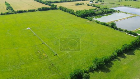Pond system and large farmland with center pivot irrigation water energy-efficient at rural Fairland, Oklahoma, pipeline mobile truss structures motorized wheels move through field, aerial view. USA