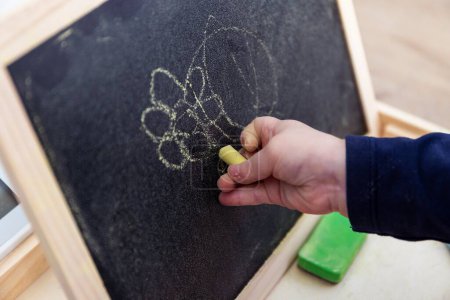 Photo for A close up portrait of a toddler drawing, scribbling or doodling a bit on a chalkboard. The child is writing on the blackboard and has a yellow piece of chalk in its hand. - Royalty Free Image