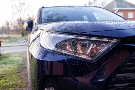 A close up portrait of the left headlight of a dark blue hybrid toyota rav4 car parked on a driveway. The vehicle can drive electrically and on fossil fuel.