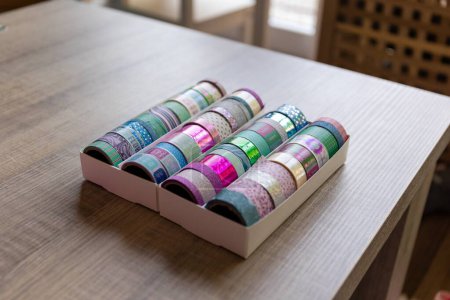 Foto de Una caja llena de rollos coloridos de cinta washi con diferentes patrones, texturas y colores en una mesa de madera. Las cintas están listas para ser utilizadas para decorar un interior, poste o tarjeta de regalo, regalo. - Imagen libre de derechos