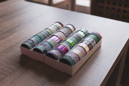 Foto de Una caja llena de rollos coloridos de cinta washi con diferentes patrones, texturas y colores en una mesa de madera. Las cintas están listas para ser usadas para decorar un poste o tarjeta de regalo, un interior, un regalo. - Imagen libre de derechos