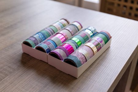 Foto de Una caja llena de rollos coloridos de cinta washi con diferentes patrones, texturas y colores en una mesa de madera. Las cintas están listas para ser utilizadas para decorar un interior, regalo o postal, regalo. - Imagen libre de derechos