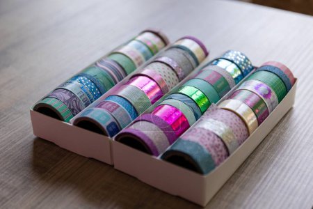 Foto de Una caja llena de rollos coloridos de cinta washi con diferentes colores, patrones y texturas en una mesa de madera. Las cintas están listas para ser usadas para decorar un poste o tarjeta de regalo, un interior, un regalo. - Imagen libre de derechos