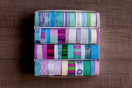 Foto de Una caja llena de rollos coloridos de cinta washi con diferentes colores, patrones y texturas en una mesa de madera. Las cintas están listas para ser utilizadas para decorar un interior, poste o tarjeta de regalo, regalo. - Imagen libre de derechos