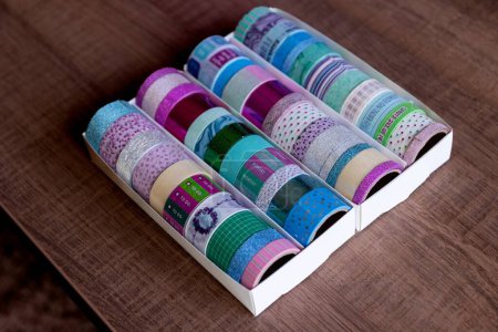 Foto de Una caja llena de rollos coloridos de cinta washi con diferentes colores, patrones y texturas en una mesa de madera. Las cintas están listas para ser usadas para decorar un regalo o postal, un interior, un regalo. - Imagen libre de derechos