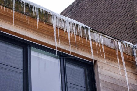 Foto de Un retrato de cerca de grandes carámbanos colgando de una alcantarilla sobre una ventana durante el invierno después de una ventisca. El hielo es peligroso y ha formado espigas afiladas. El techo está cubierto de nieve blanca. - Imagen libre de derechos